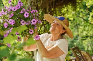 Happy senior woman tends the flowers in a hanging pot. There is a green background of blurred plants, and wooden outdoor chairs are visible in the lower right corner.